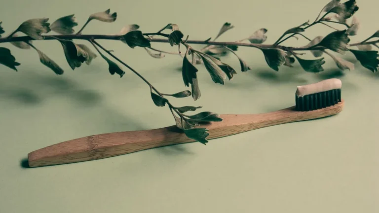 A bamboo toothbrush with toothpaste lies on a green background adorned with leaves, promoting natural oral care.