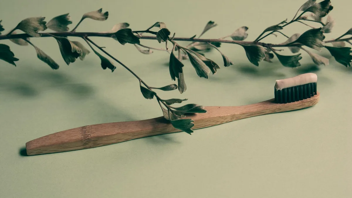 A bamboo toothbrush with toothpaste lies on a green background adorned with leaves, promoting natural oral care.