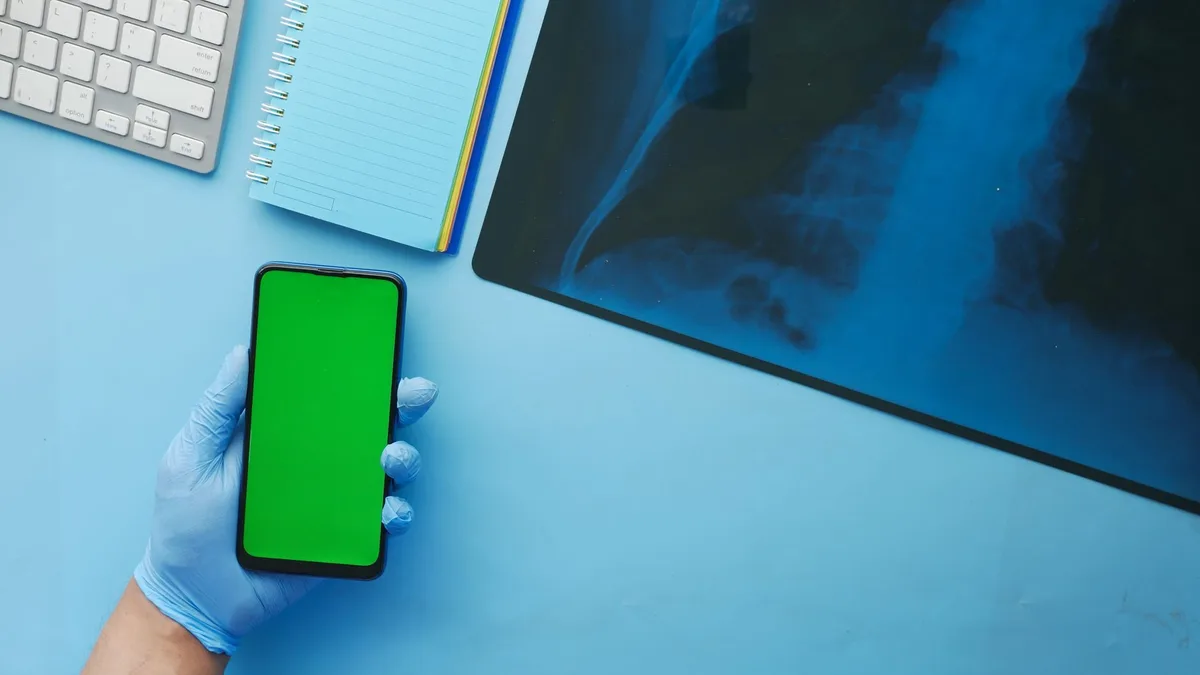 Close-up of a hand in a glove holding a smartphone with a green screen next to x-ray on a blue desk.